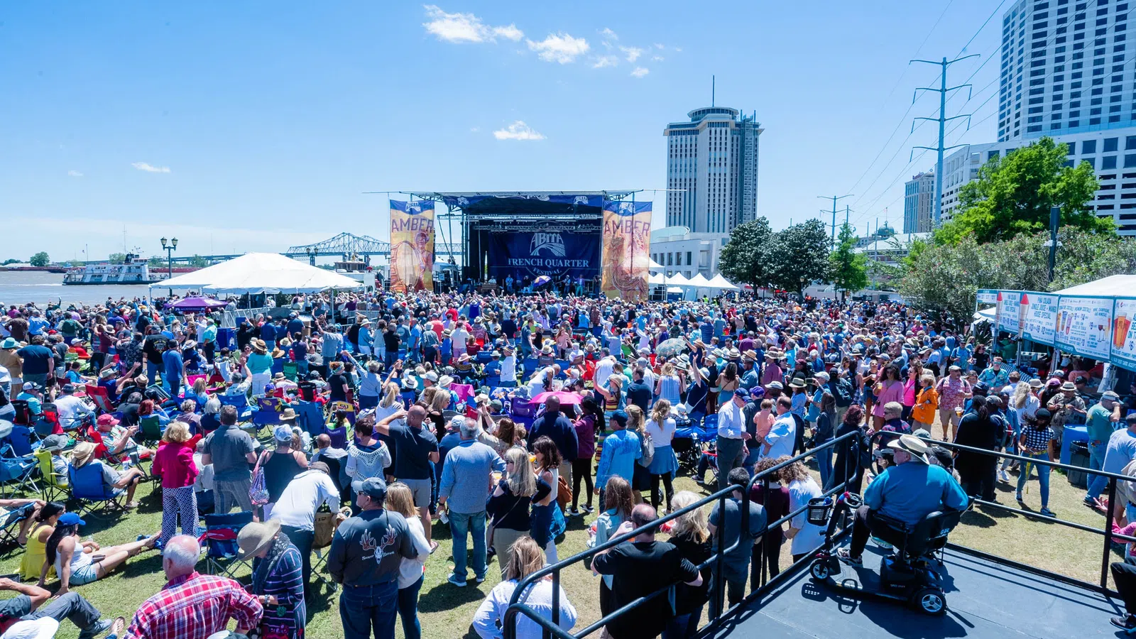Large crowd gathered outdoors at a sunlit music festival near a river with stage and city skyline in the background