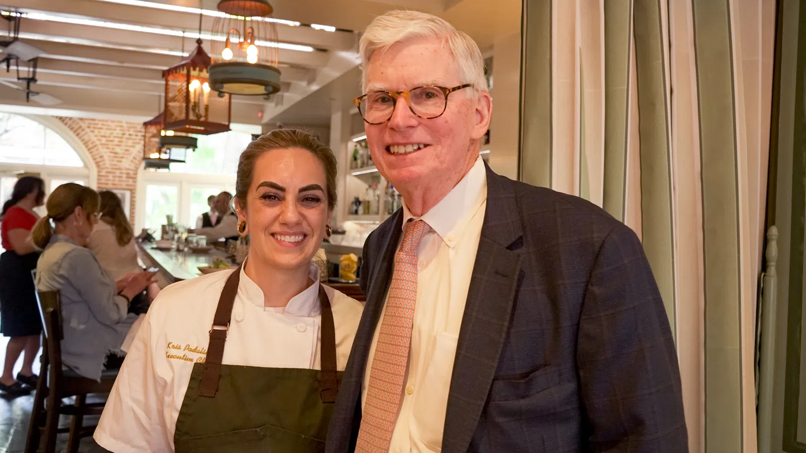Chef and man in suit smiling inside a warmly lit restaurant with patrons at the bar in the background