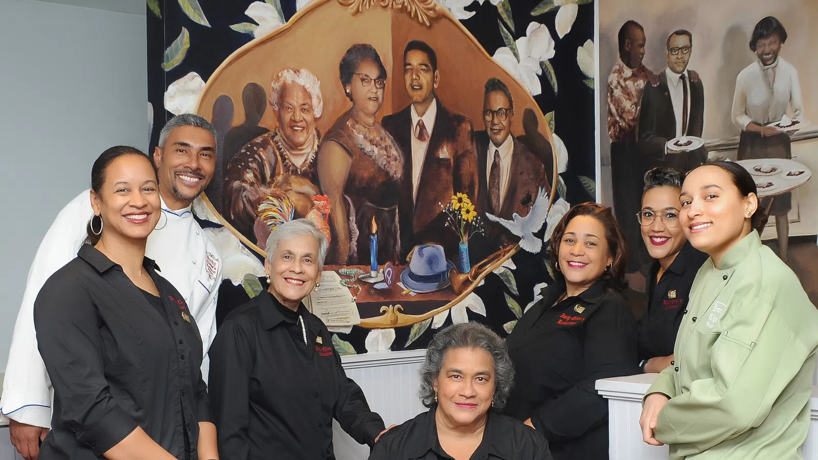 Group of diverse restaurant staff posing inside with a mural of historical figures behind them