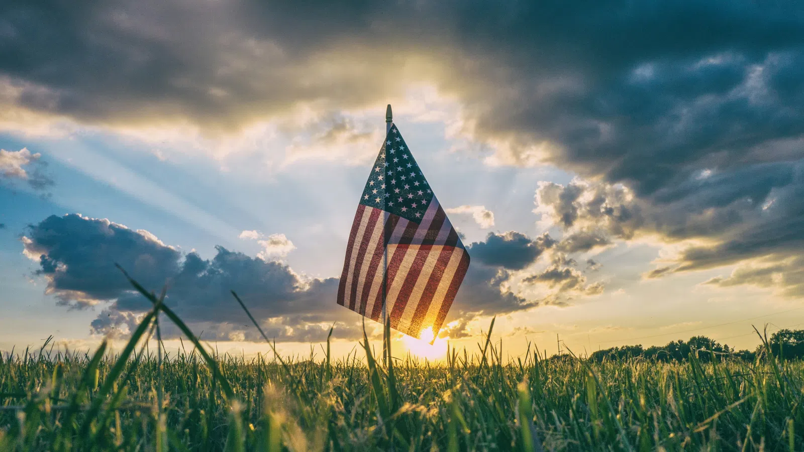 American flag standing in a grassy field with sun rays and dramatic cloudy sky at sunset