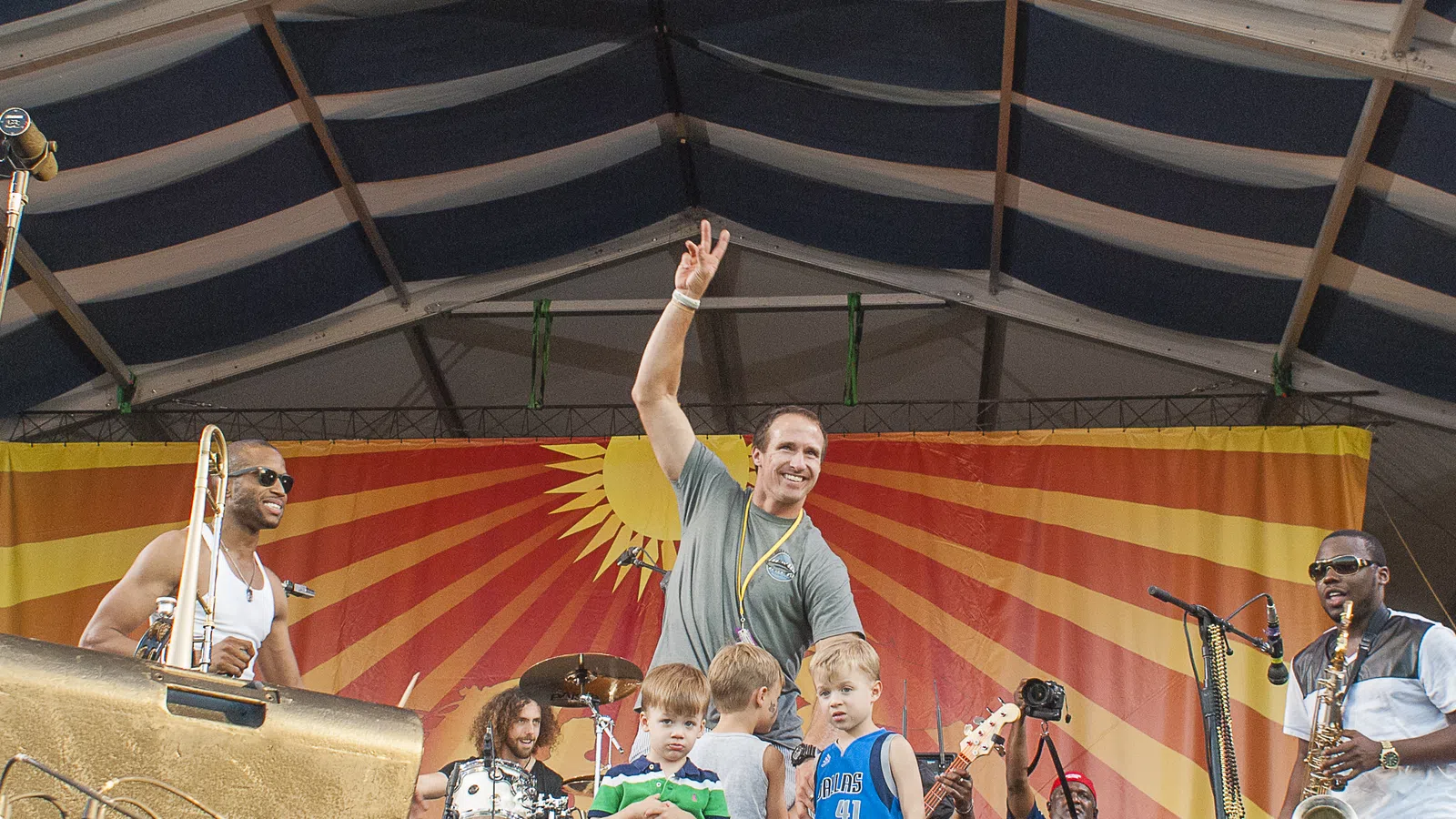 Musicians and children on stage at outdoor concert with colorful sunburst backdrop under a tent roof