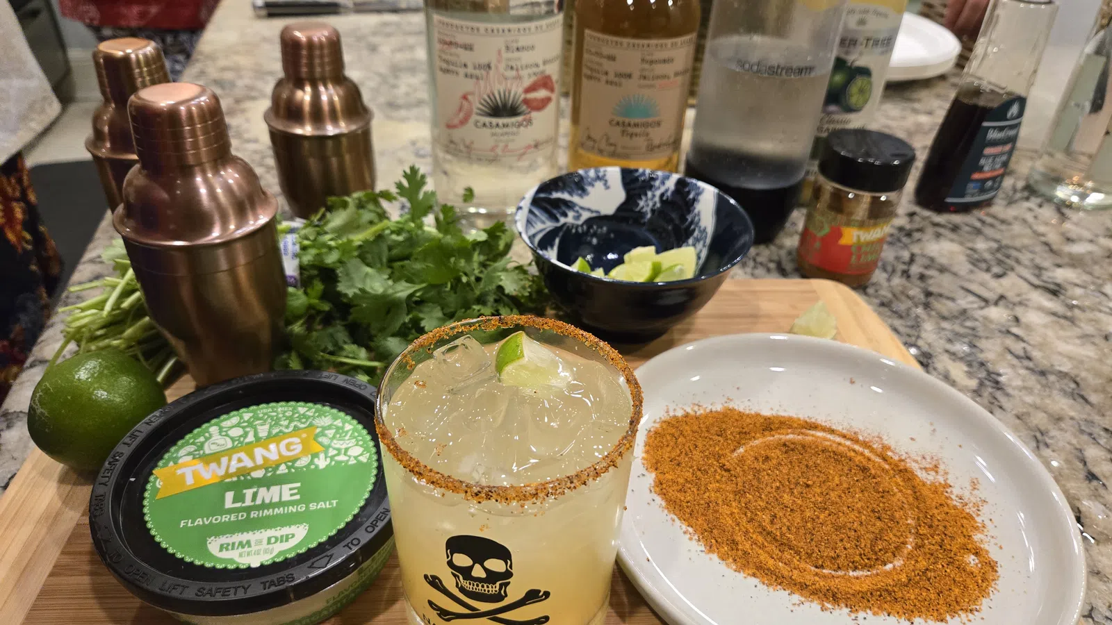 Margarita cocktail with chili rim, lime, seasoning salt, cilantro, and cocktail mixers on wooden board.