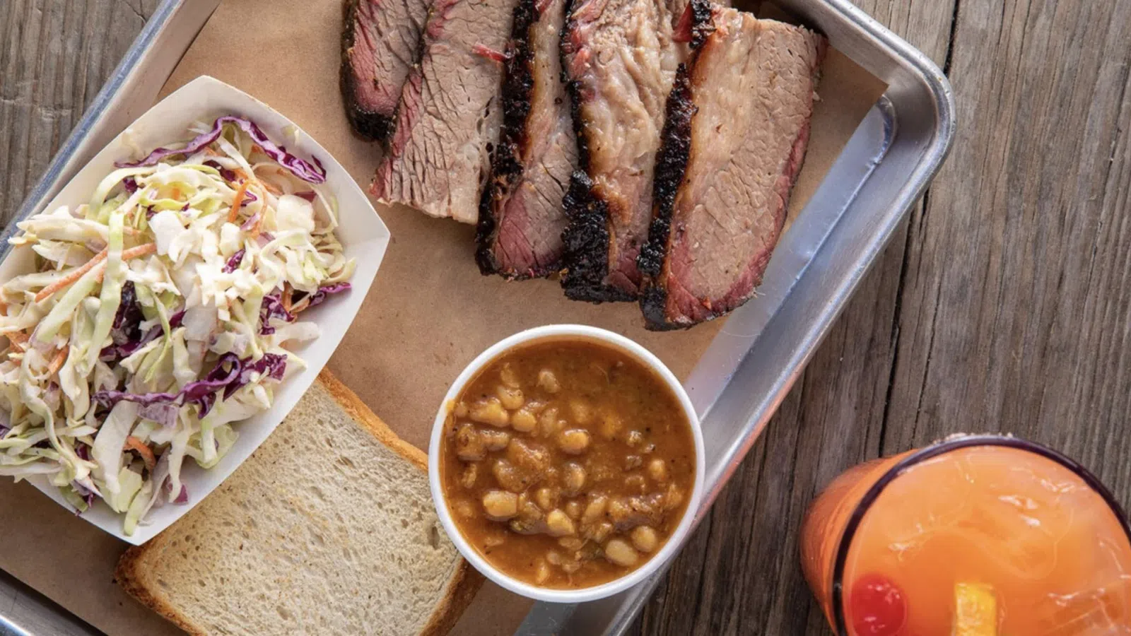BBQ platter with sliced smoked brisket, baked beans, coleslaw, white bread, and a cocktail on a wooden table.