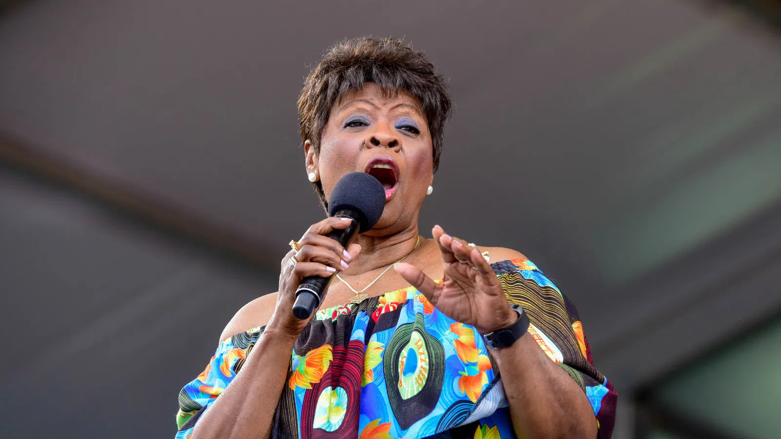Female singer performing passionately on stage wearing a colorful off-shoulder floral dress and holding a microphone.
