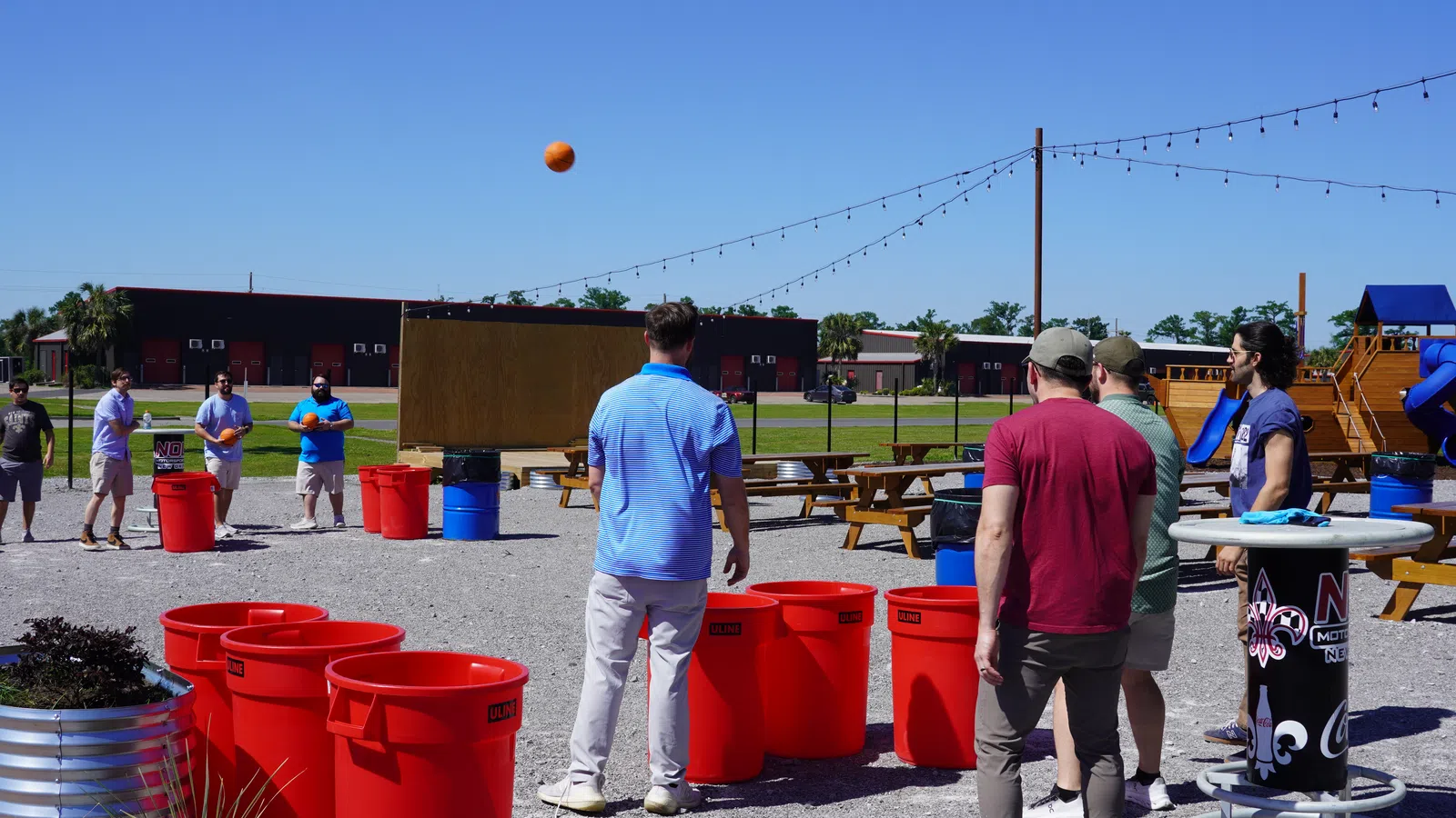 Group of men playing outdoor game tossing balls into large red bins on a sunny day with picnic tables and playground nearby