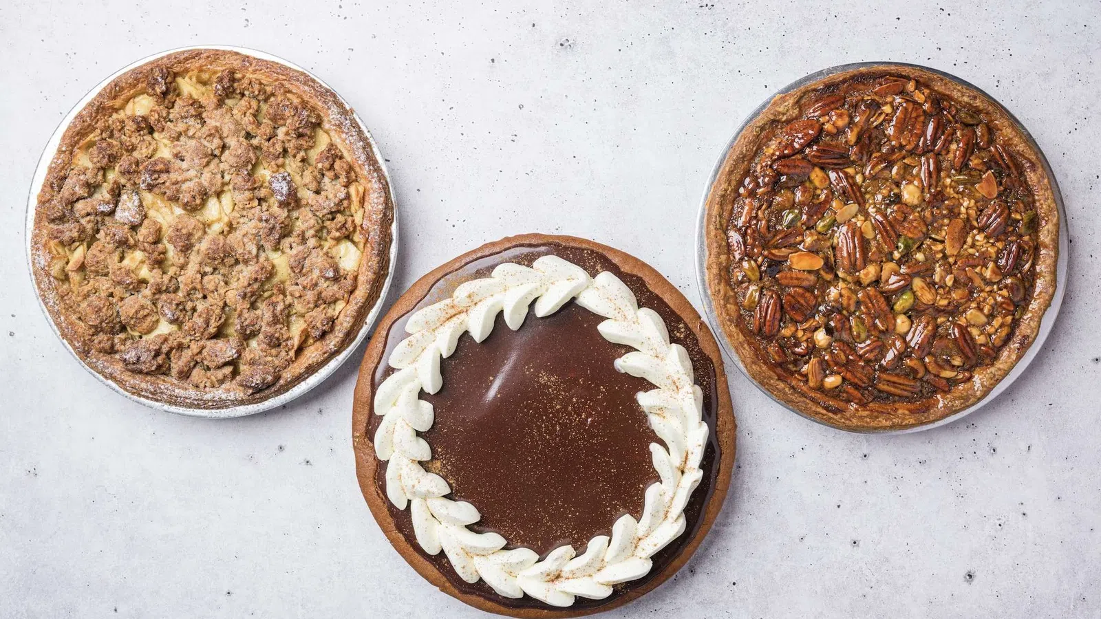 Three round pies displayed on a light surface: apple crumb, chocolate with whipped cream, and mixed nut pecan pie.