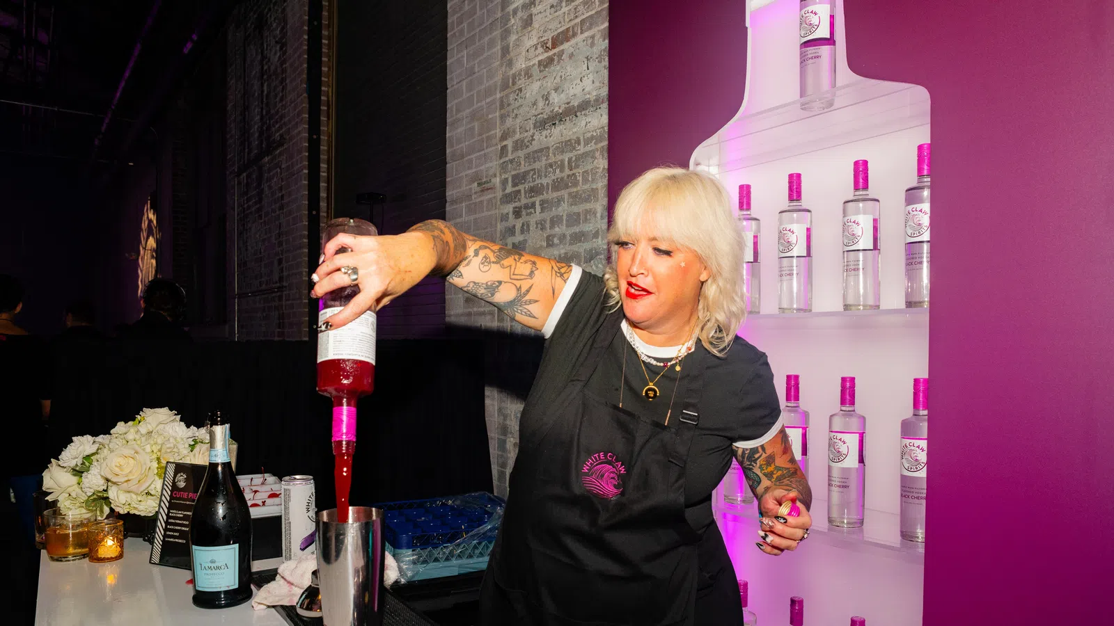 Bartender with tattoos pouring pink liquid into shaker at bar with bottles and flowers in background