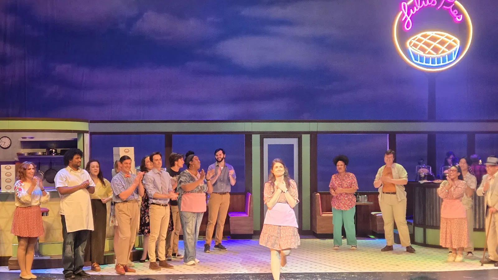 Cast of a theatrical play standing on stage with a diner set and neon pie sign in the background.