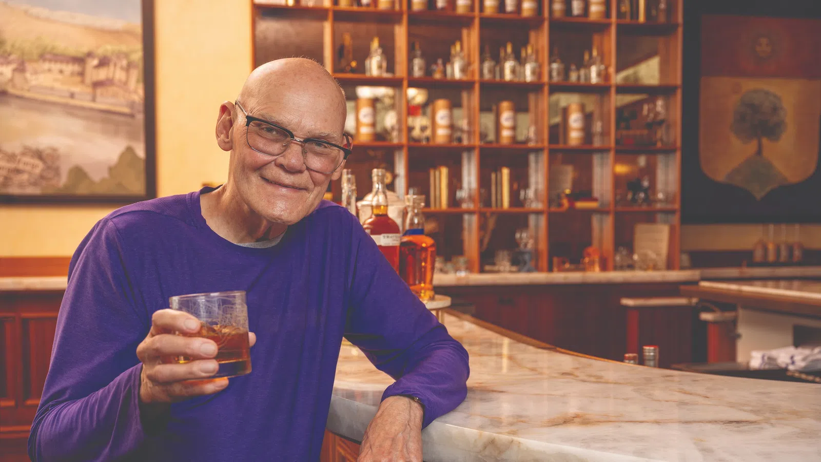 Elderly man in purple shirt sitting at bar holding glass of whiskey with shelves of liquor in background