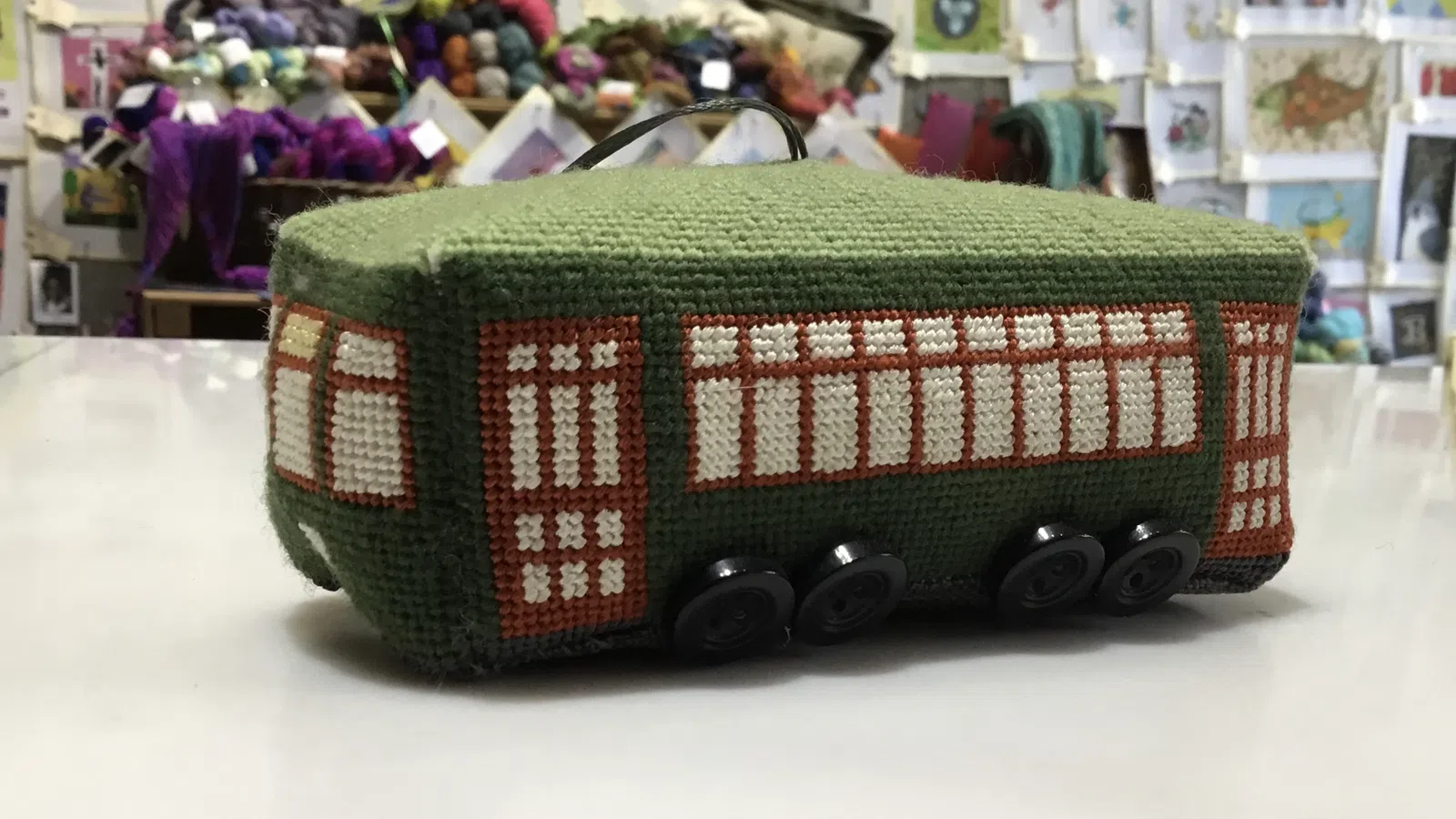 Handcrafted green knitted bus with window patterns and button wheels displayed on a white table in a craft store.