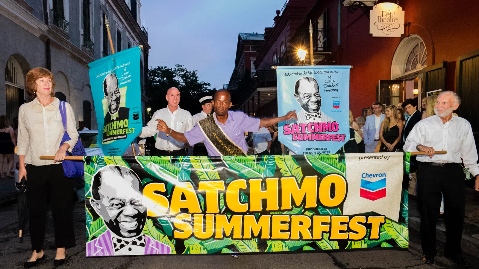 People holding a colorful Satchmo SummerFest banner during an evening street event with buildings and onlookers.