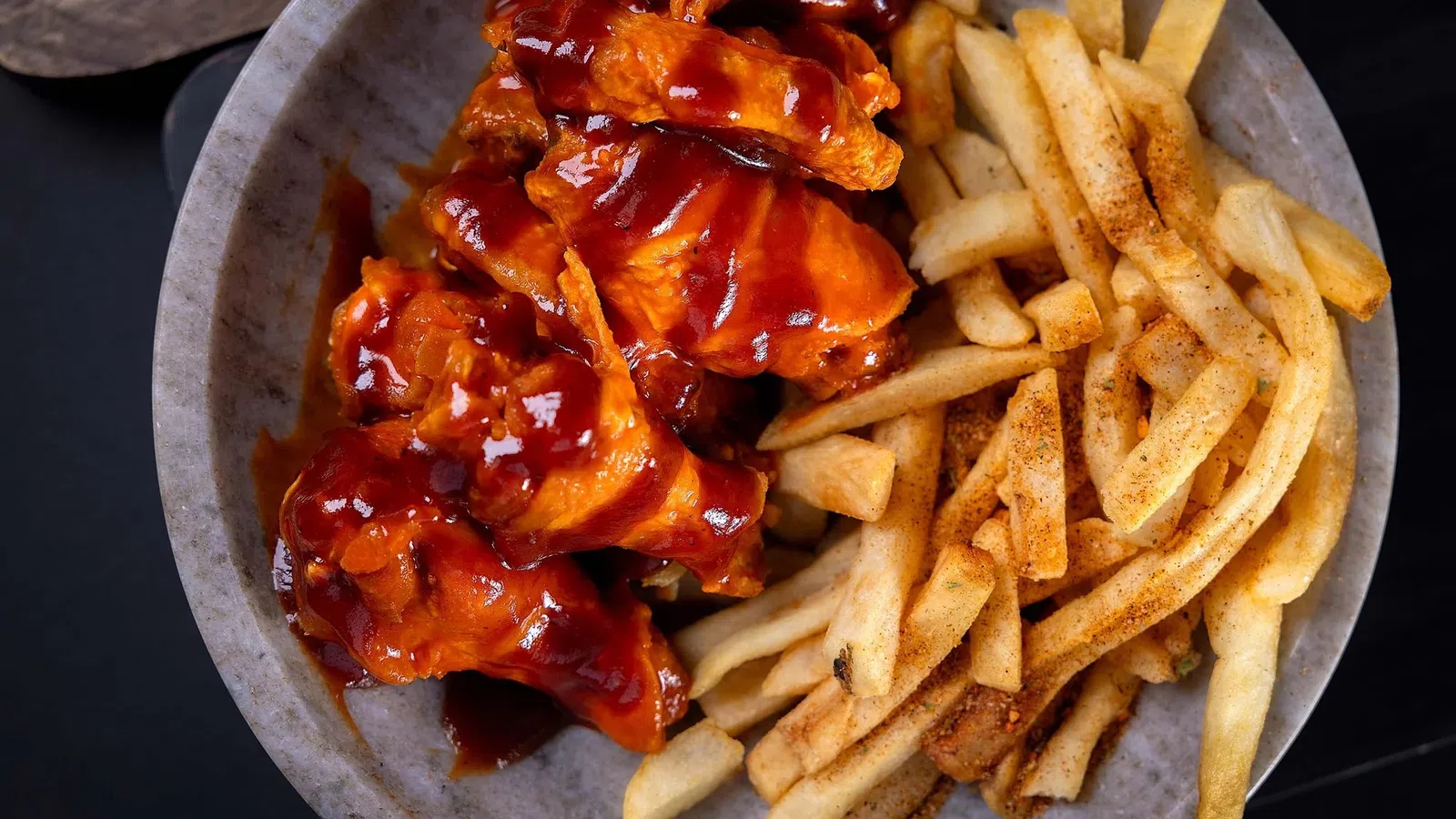 Plate of barbecue chicken wings and seasoned French fries on a gray dish with dark background.