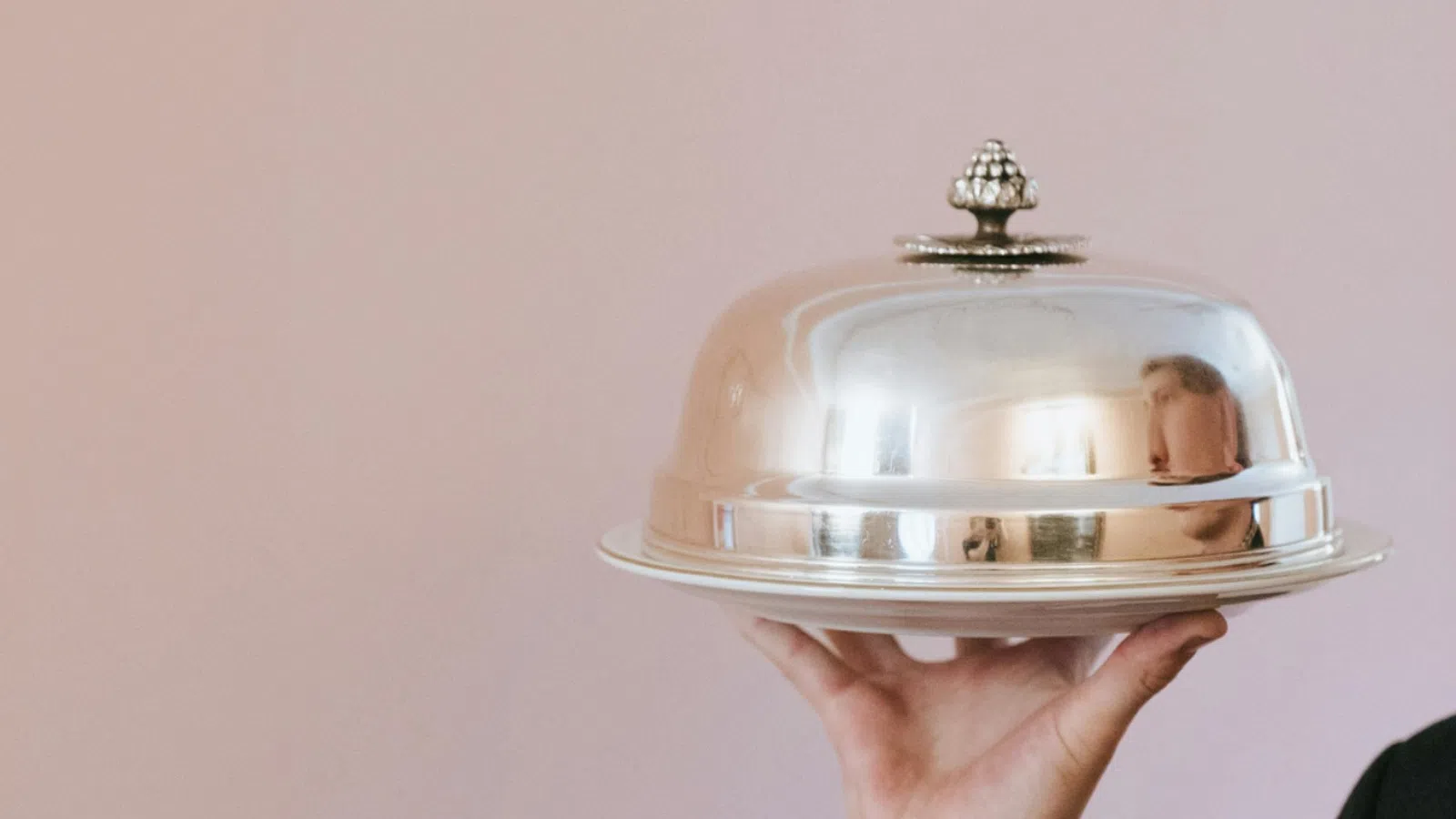 Person holding a silver cloche food cover on a tray against a soft pink background