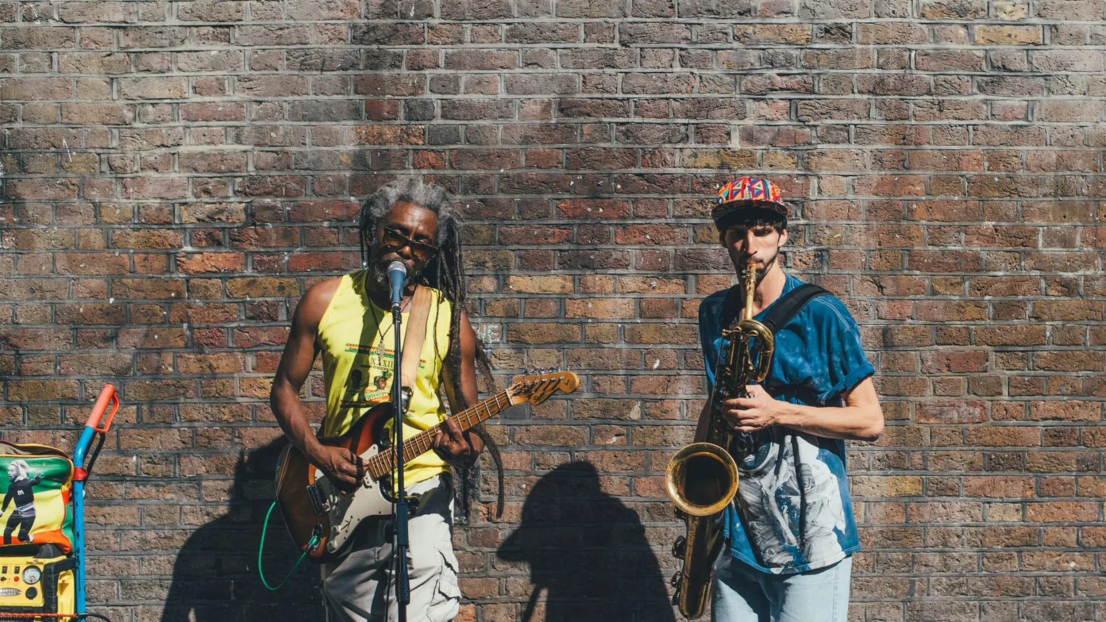 Two street musicians perform against a brick wall, one playing electric guitar and singing, the other playing saxophone.