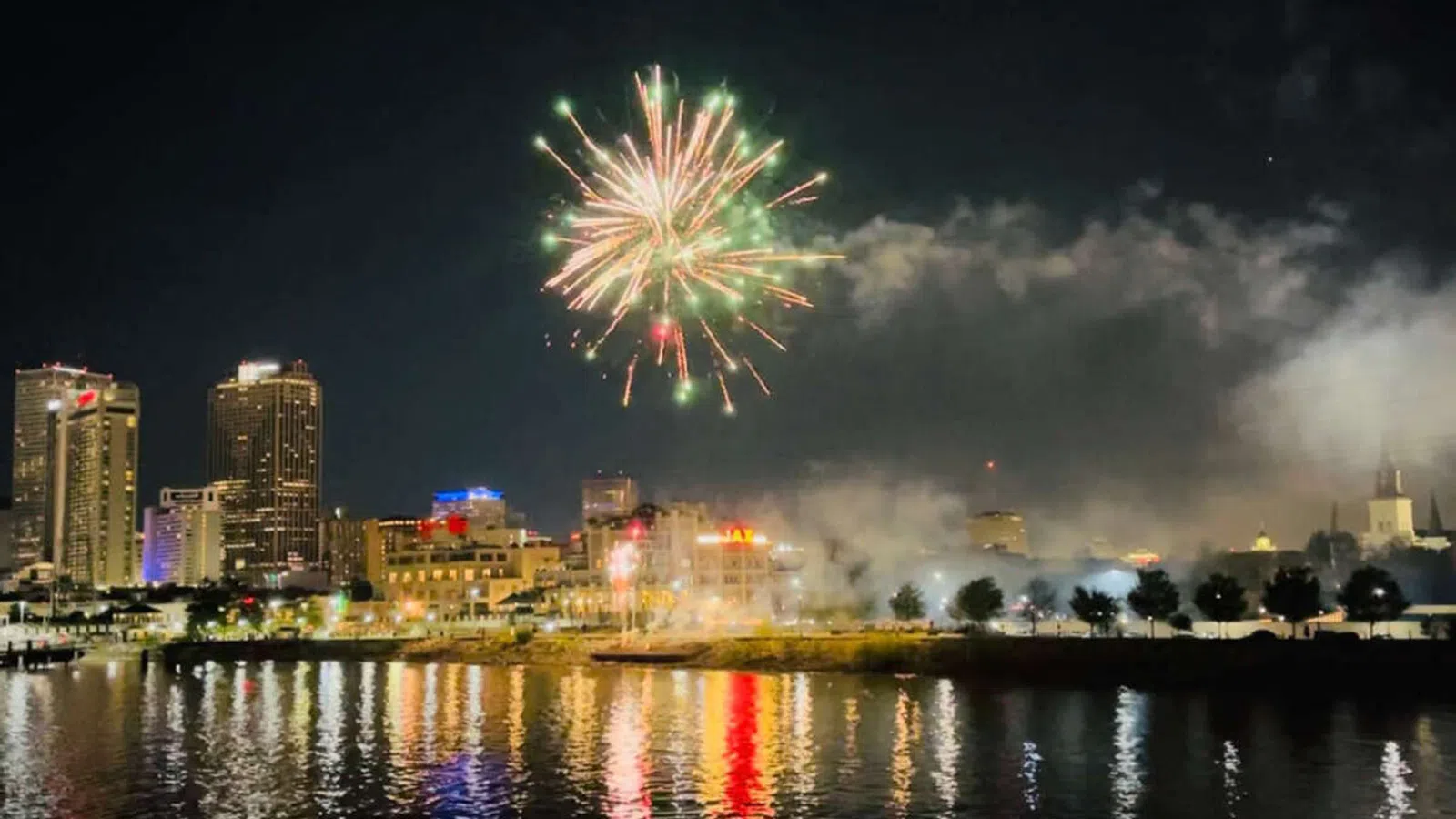 Colorful fireworks burst above a city skyline at night reflecting on calm river water below.