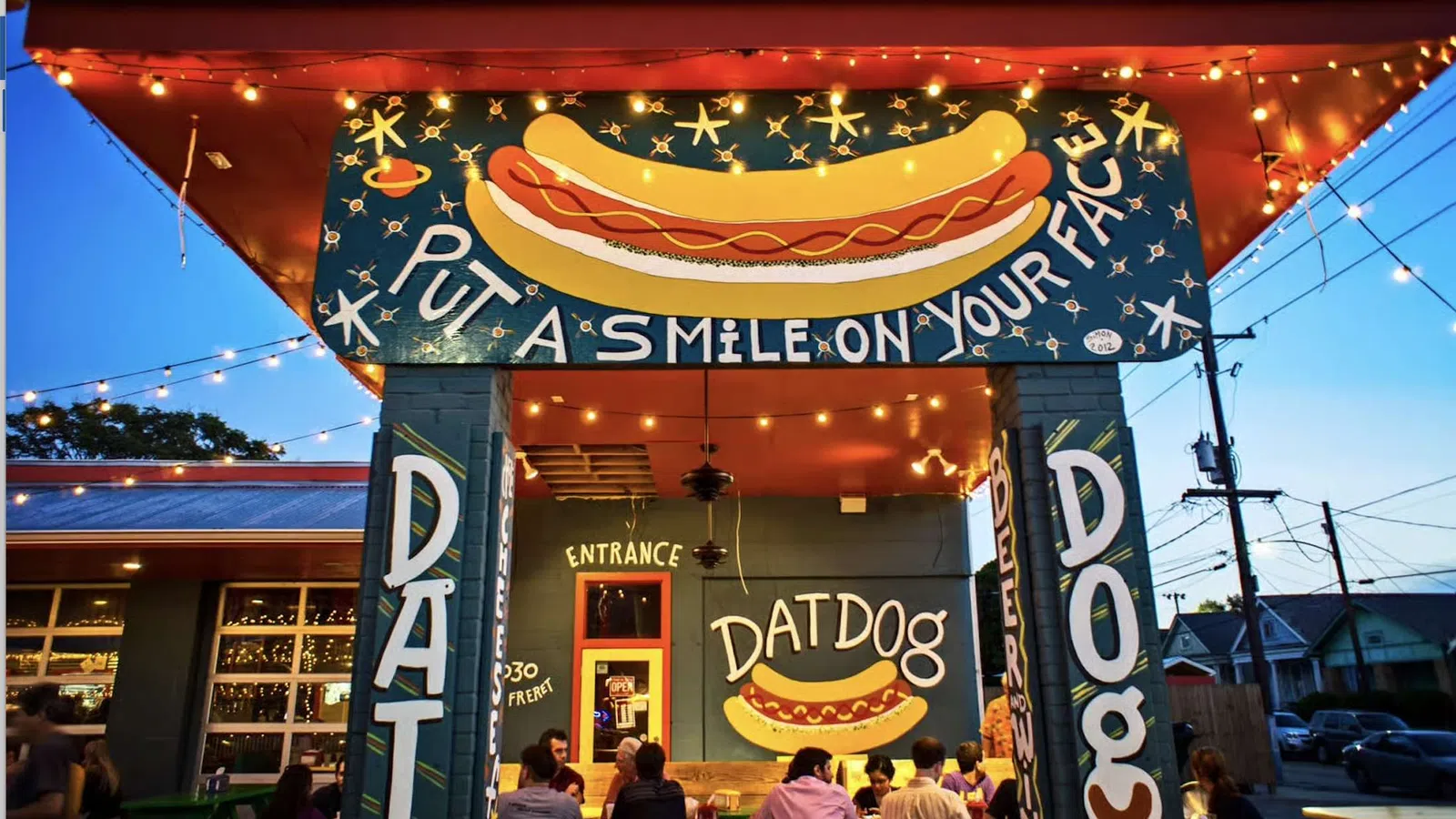 Front entrance of Dat Dog restaurant with colorful hot dog sign and dining patrons at dusk under string lights