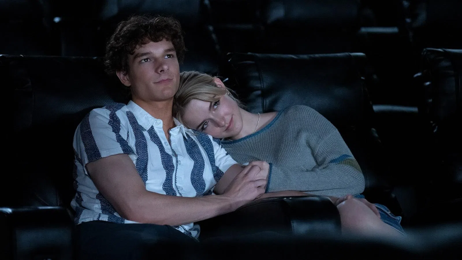 Young couple sitting closely in a dark movie theater, woman resting her head on man's shoulder holding hands.