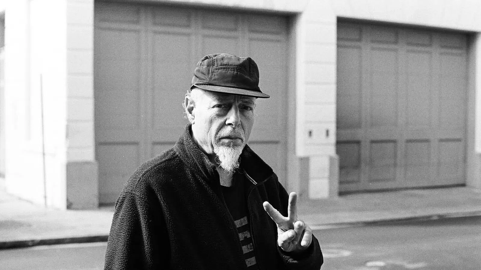 Black and white photo of a man with a goatee wearing a cap and jacket showing a peace sign outdoors on street.