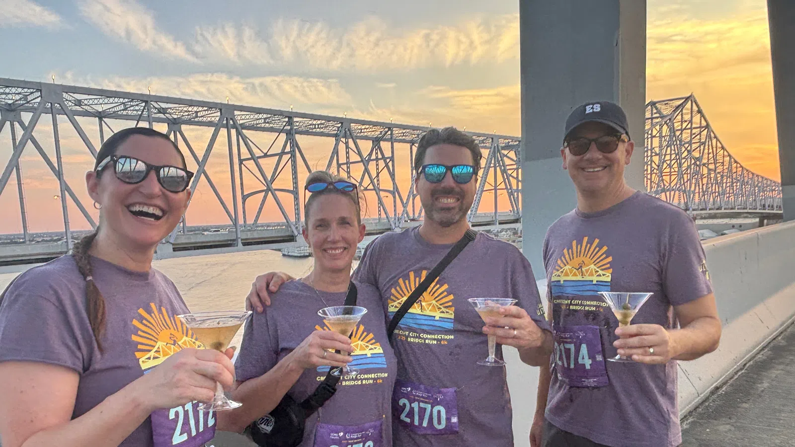 Four runners wearing matching purple shirts hold drinks by a bridge at sunset celebrating their run.