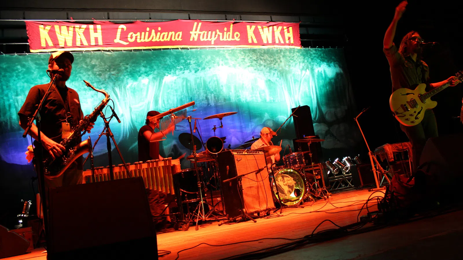 Live band performing on stage under KWKH Louisiana Hayride banner with saxophone, drums, and guitar.