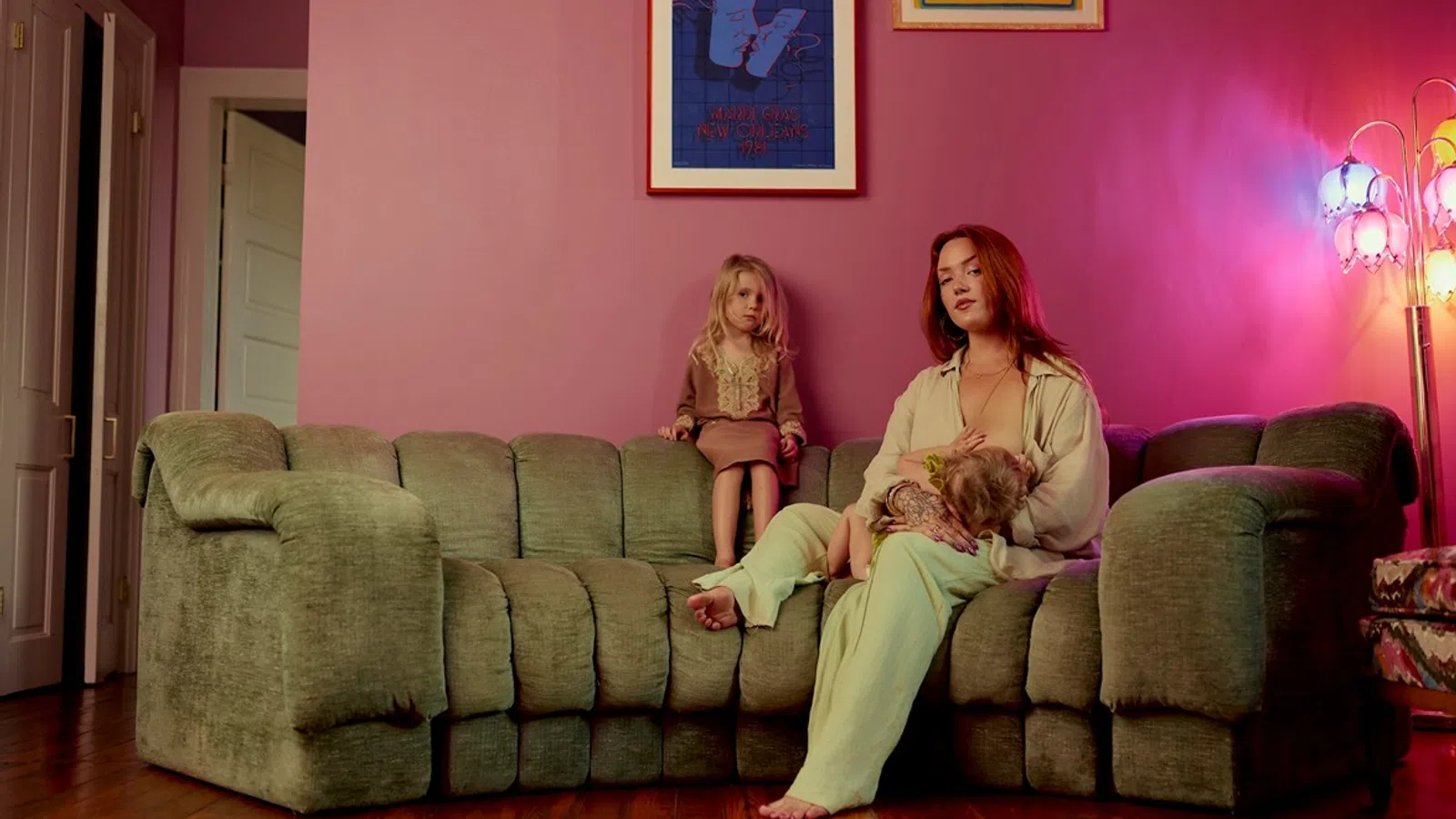 Mother breastfeeding baby on green couch while young girl sits beside them in pink room with framed art.