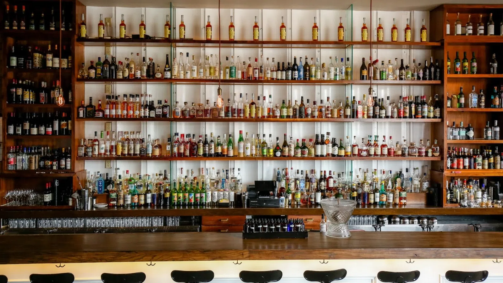 Wide bar with rows of liquor bottles on shelves and empty black bar stools lined up at wooden counter.