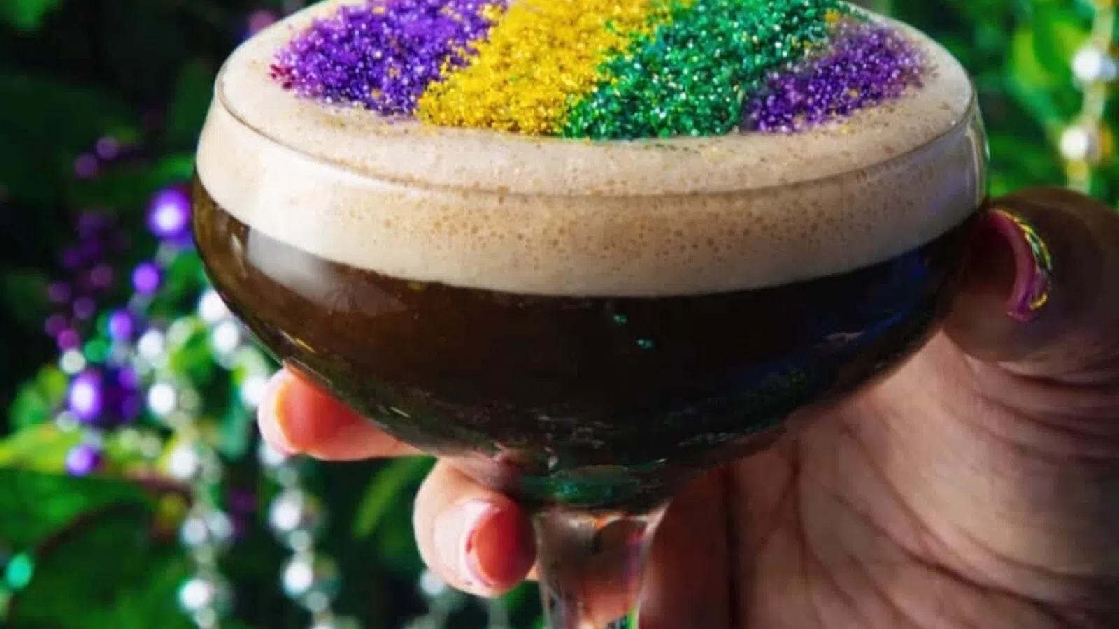 Hand holding a cocktail glass with dark drink topped with purple, yellow, and green glitter foam for Mardi Gras celebration.