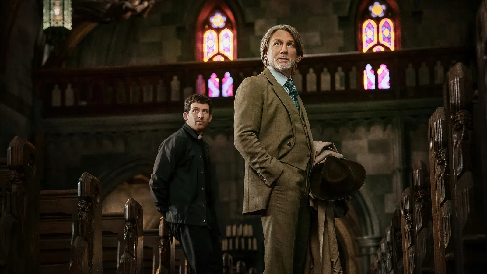 Two men, one in a suit holding a hat, standing inside a church with stained glass windows and wooden pews.