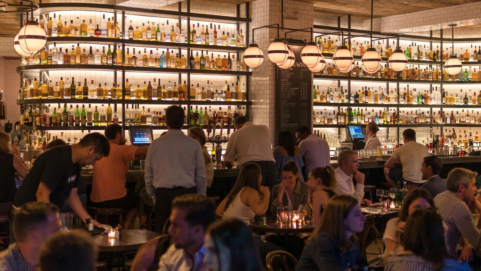 Busy bar scene with patrons sitting and standing, illuminated bottles on shelves, and hanging globe lights.