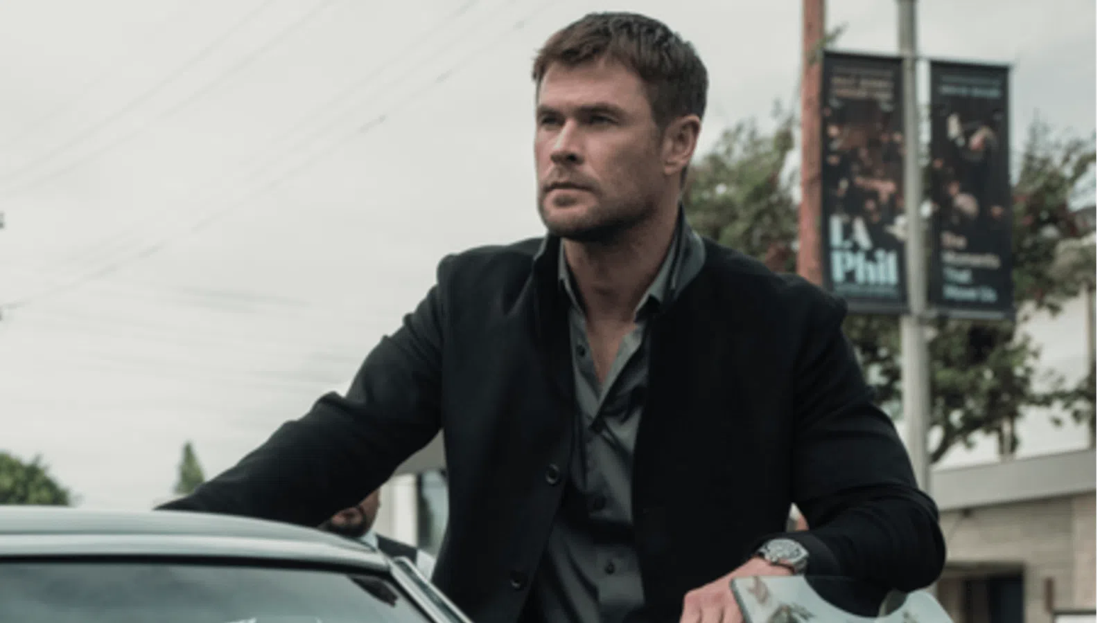 Man in black jacket leaning on a vintage car in an urban street setting with utility poles in the background.