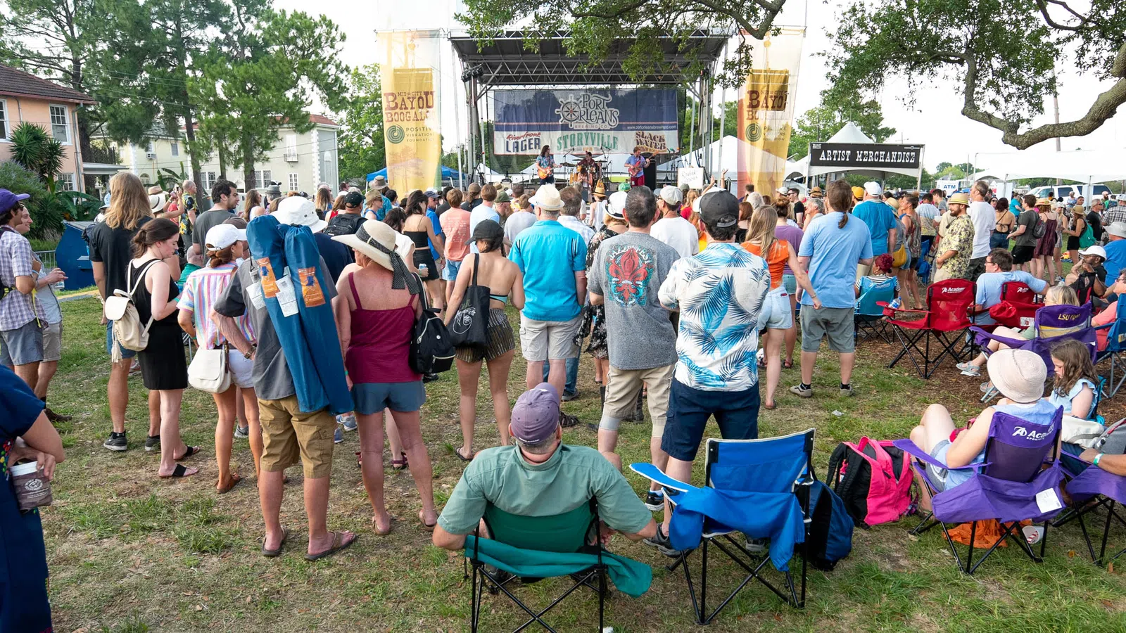 Crowd of people enjoying outdoor music festival with stage, banners, and attendees in casual summer attire.