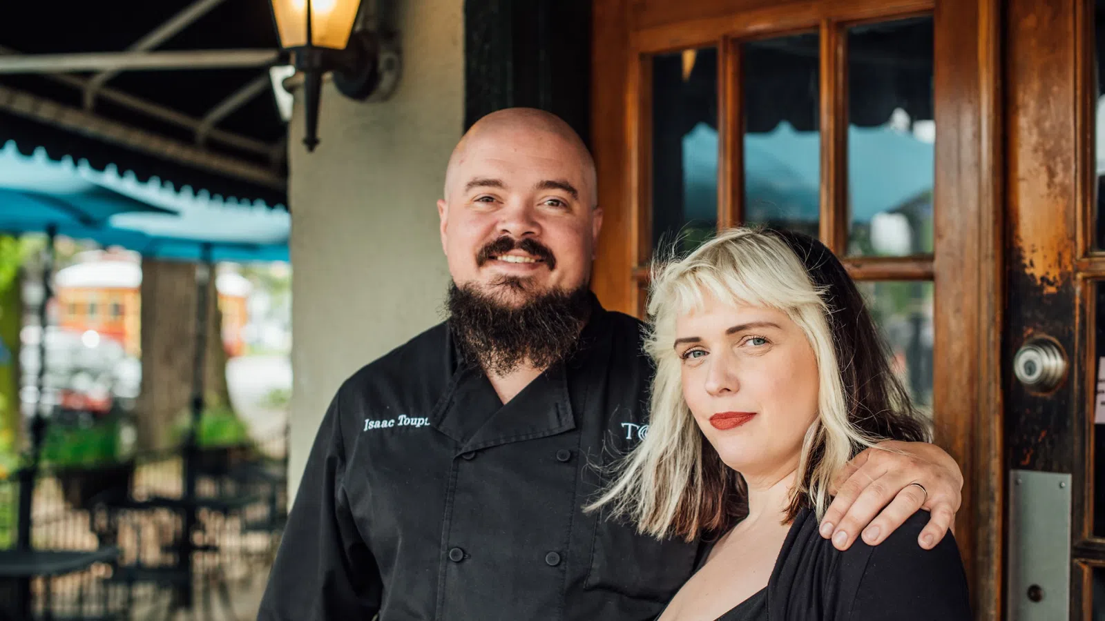 Smiling chef with beard in black uniform embraces woman with blonde hair outside restaurant entrance.
