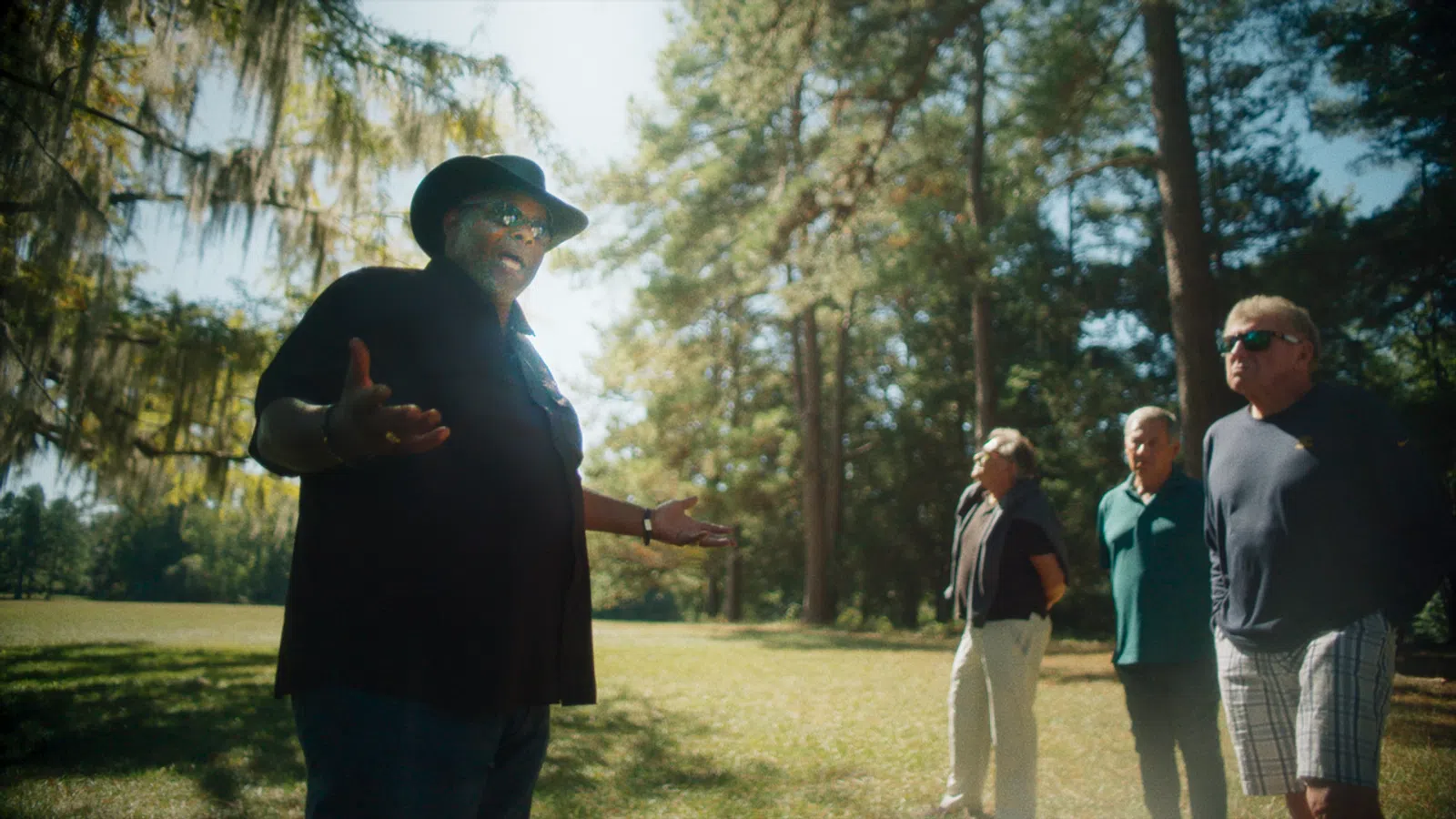 Man in black hat and shirt speaking to three men standing in a sunlit forest clearing.