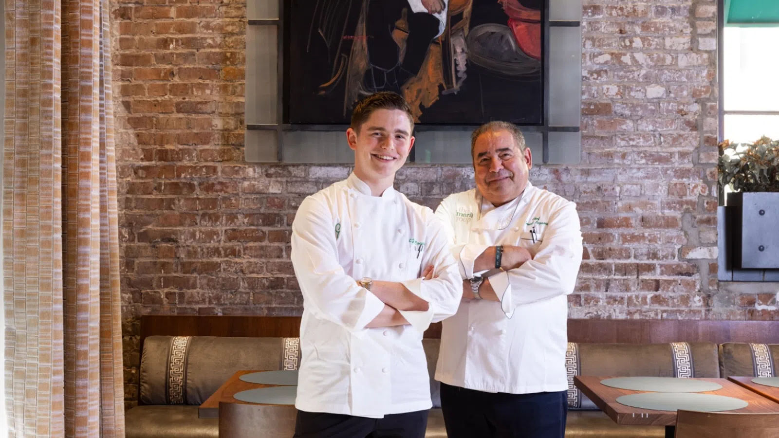 Two chefs in white uniforms standing in a rustic restaurant with brick walls and a chef portrait painting.