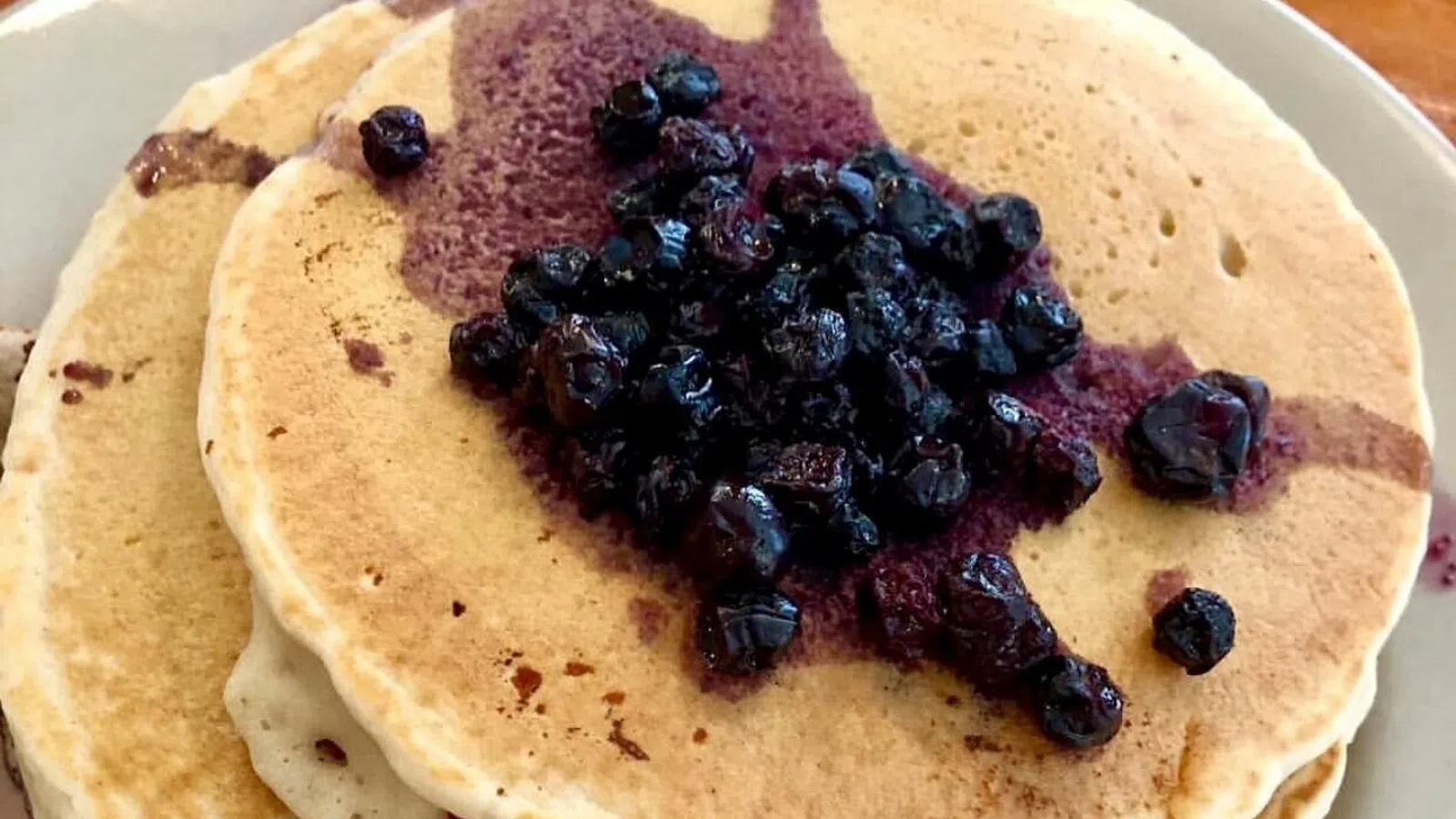 Stack of golden brown pancakes topped with juicy cooked blueberries and blueberry syrup on a plate.