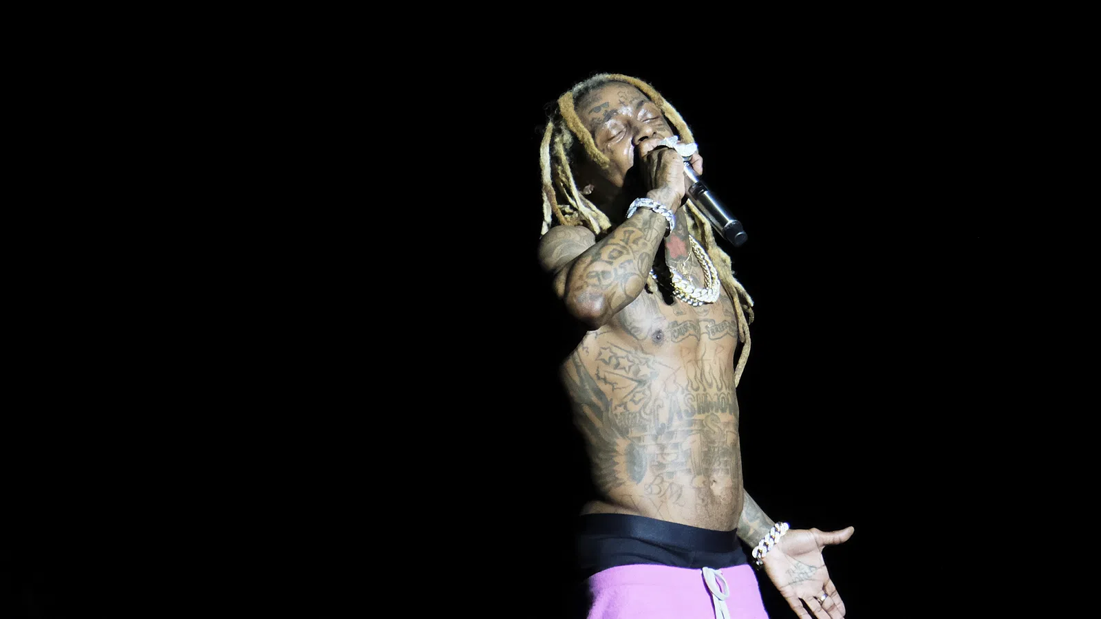 A tattooed rapper performs passionately on stage with a microphone, spotlighted against a dark background.