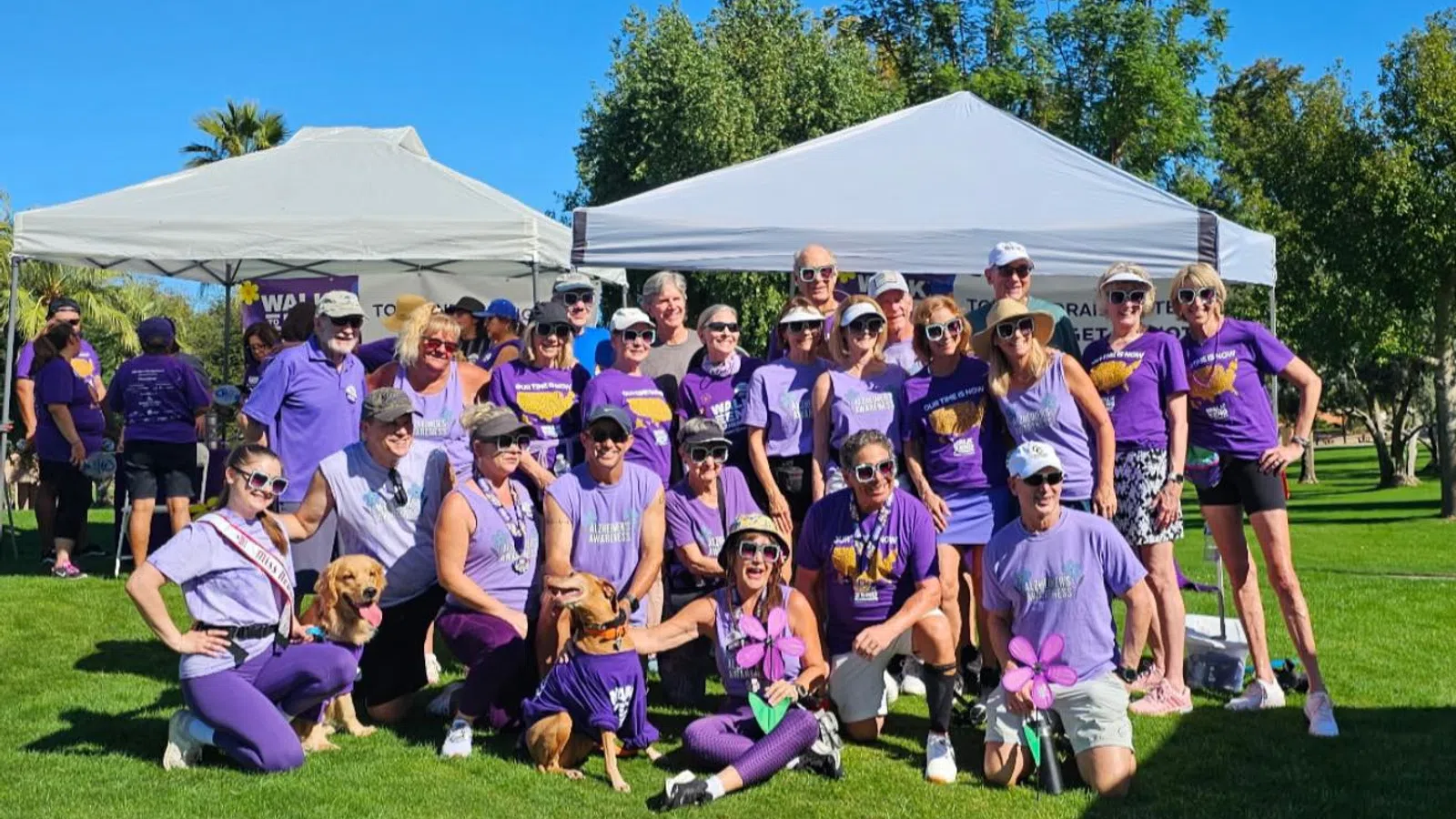 Group of people wearing purple shirts posing outdoors with two dogs under white tents on green grass