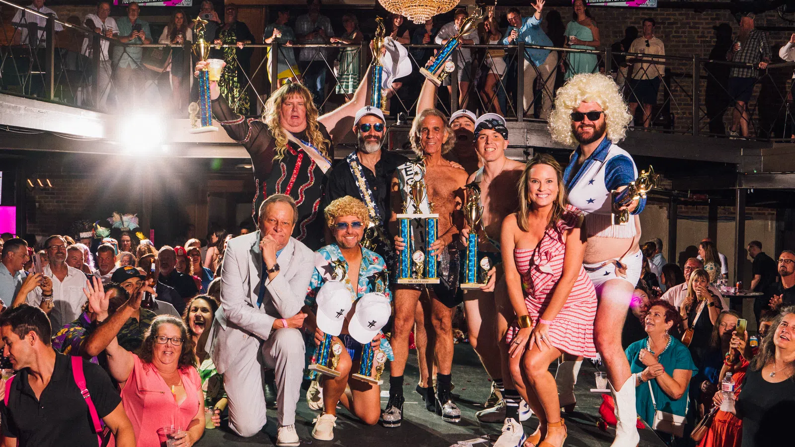Group of people in colorful costumes holding trophies on stage with cheering audience and chandelier overhead