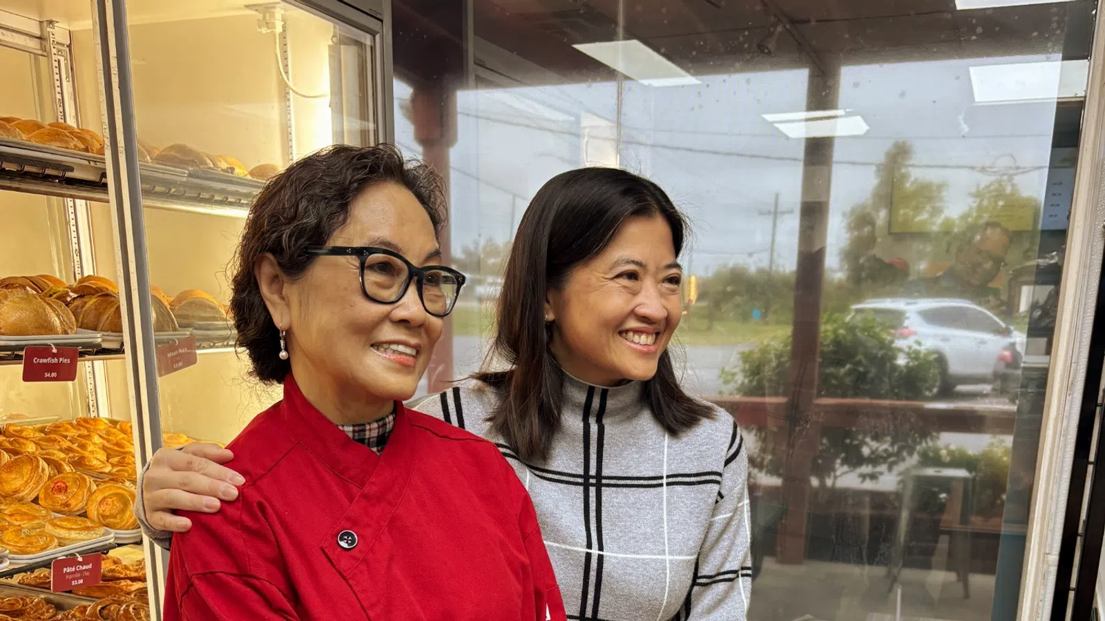 Two women smiling inside a bakery, one wearing a red coat and glasses, the other in a gray plaid sweater.