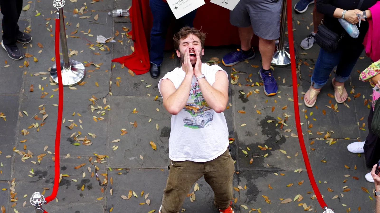 Man inside red ropes shouting or calling out while onlookers stand outside on a leaf-strewn pavement.