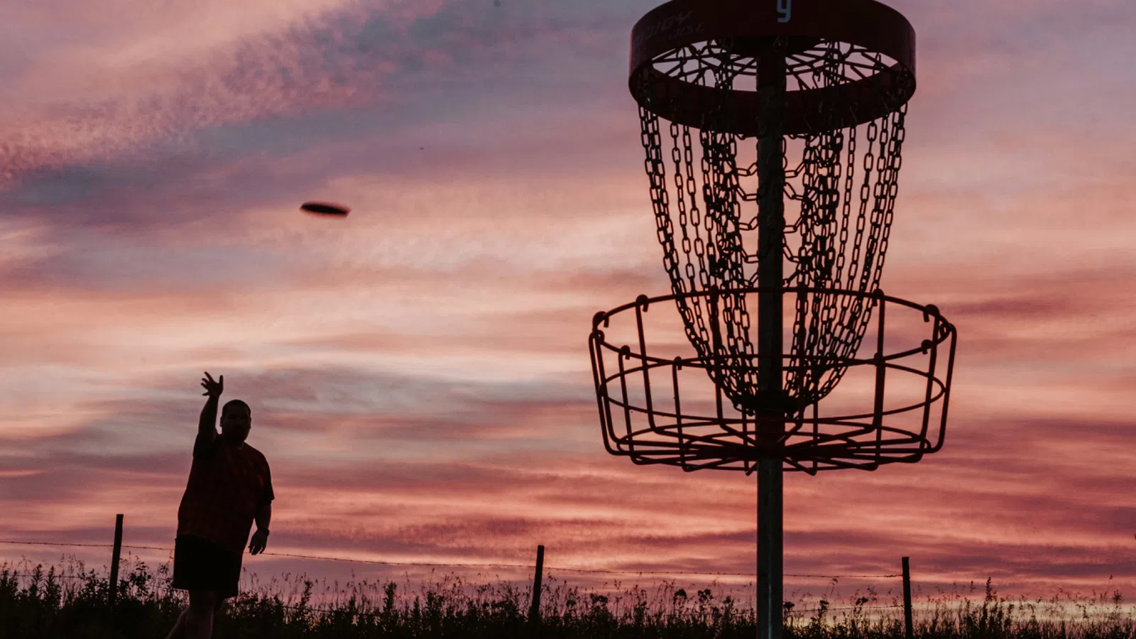 Silhouette of person throwing disc towards disc golf basket at sunset with colorful sky