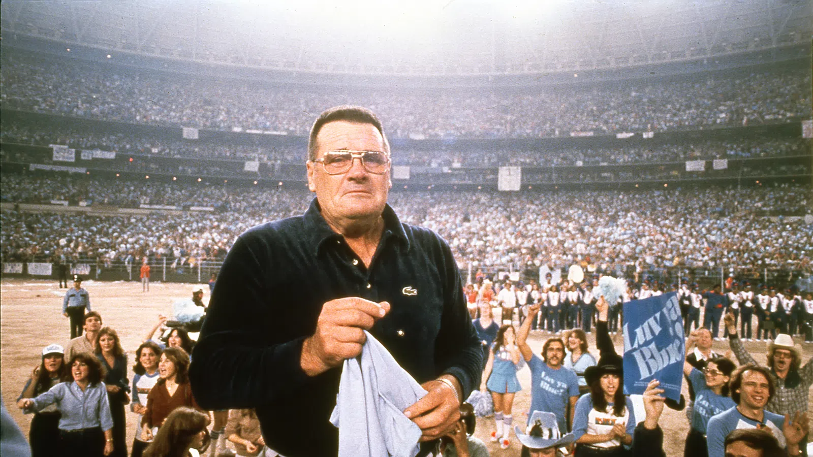 Man in glasses holding a towel at a crowded stadium event with cheering fans in the background