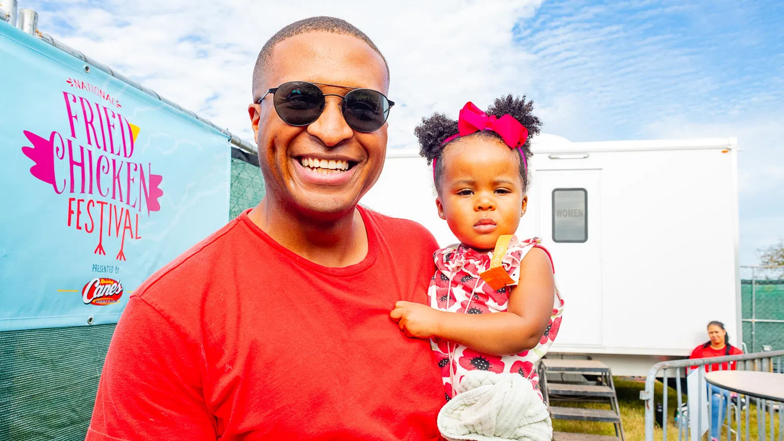 Smiling man in sunglasses holding toddler girl with bow at outdoor fried chicken festival under blue sky