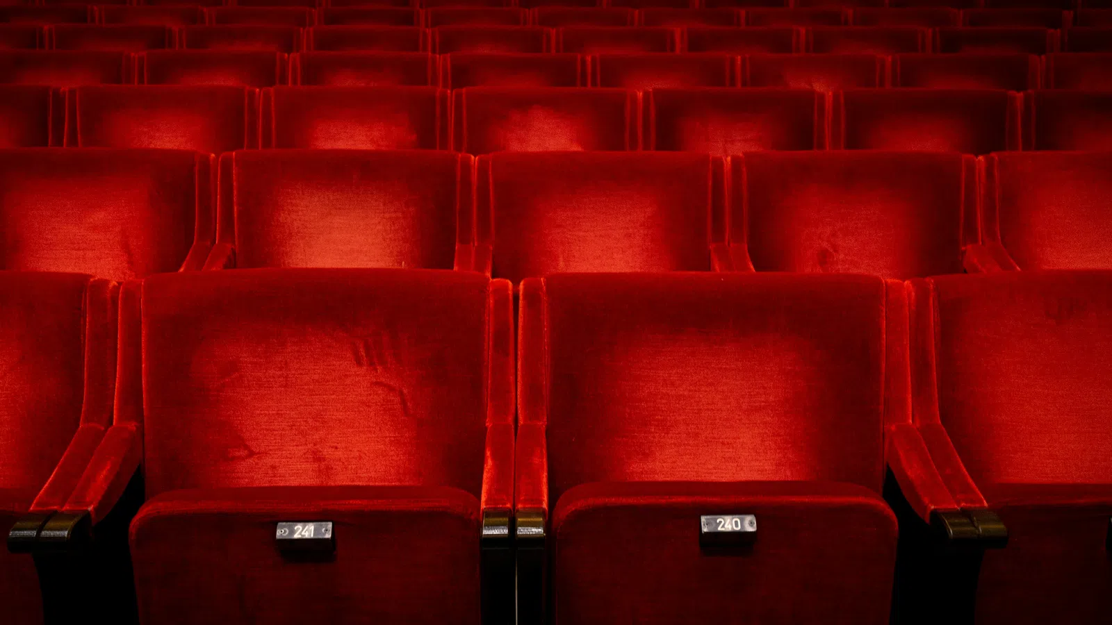 Empty red velvet theater or cinema seats arranged in rows with seat numbers visible on each seat.