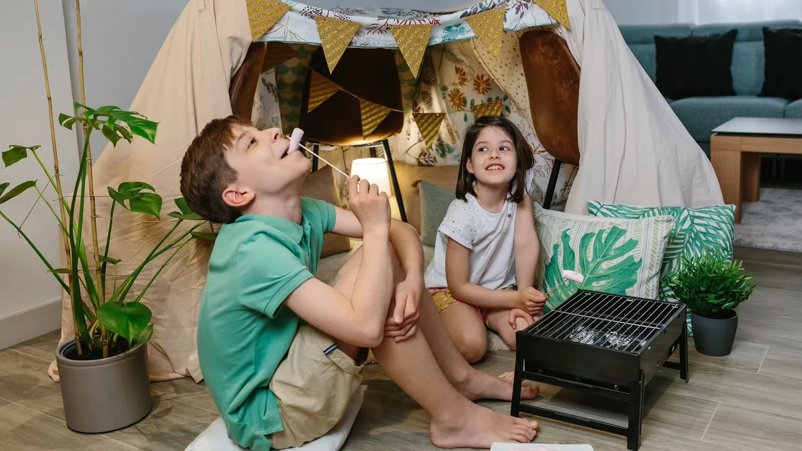 Two children enjoying indoor camping with a blanket tent and roasting marshmallows on a small grill.