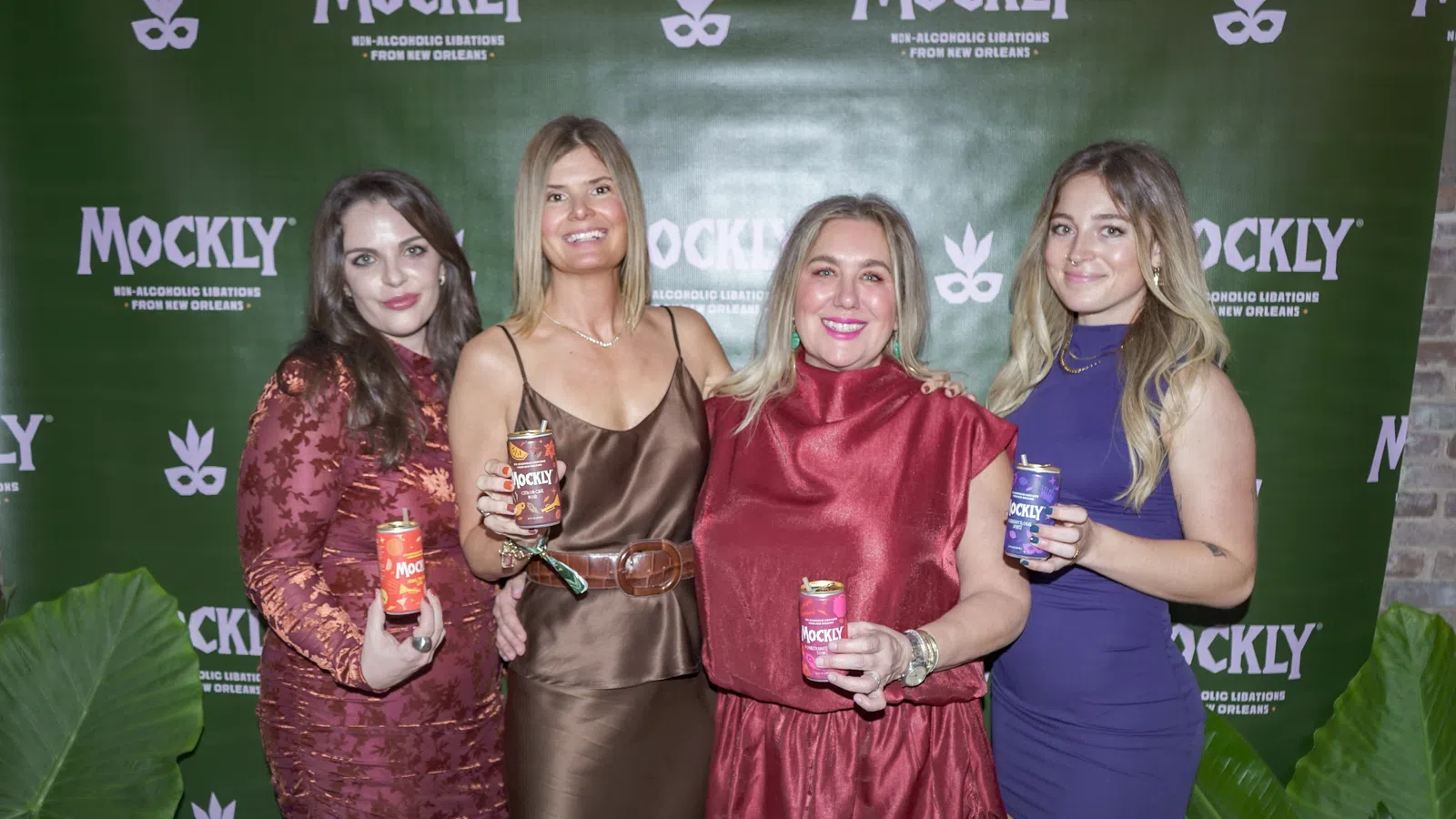 Four women posing with Mockly non-alcoholic beverage cans in front of a branded green backdrop at an event.