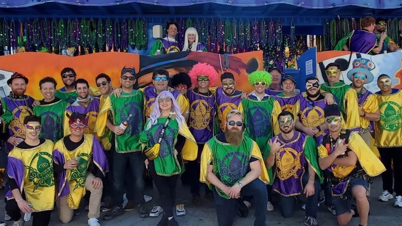 Group of people wearing colorful Mardi Gras costumes and wigs posing in front of festive decorations outdoors.
