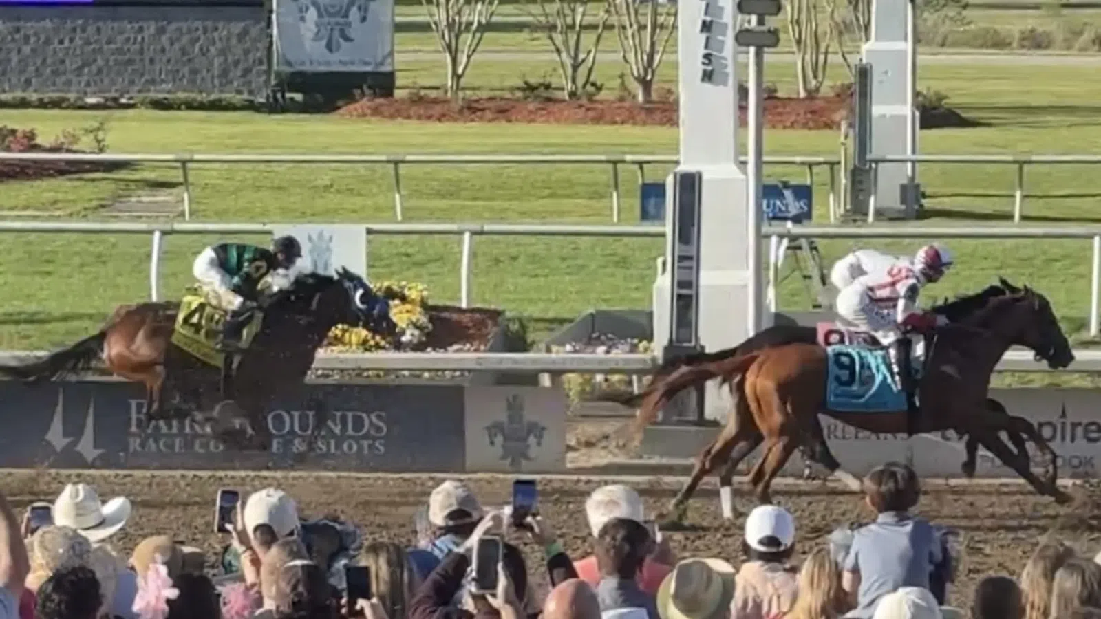 Two racehorses sprinting to the finish line as a large crowd watches and records with phones at a racetrack.