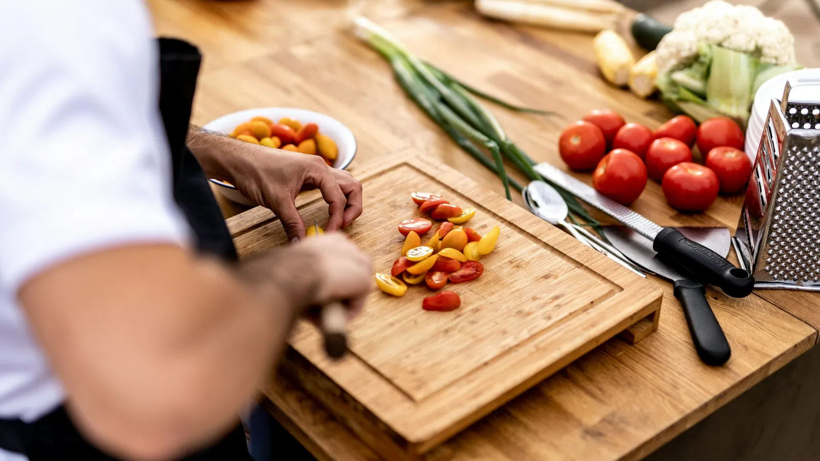 Person chopping red and yellow cherry tomatoes on a wooden cutting board with fresh vegetables nearby