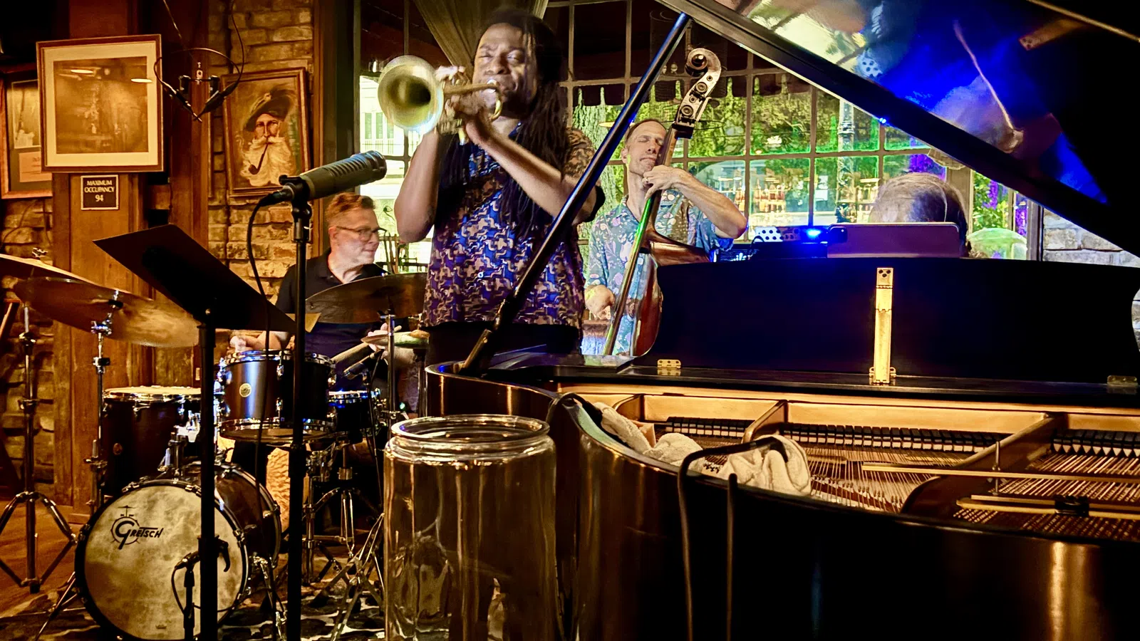Jazz band performing live with trumpet, drums, double bass, and grand piano in cozy indoor venue.
